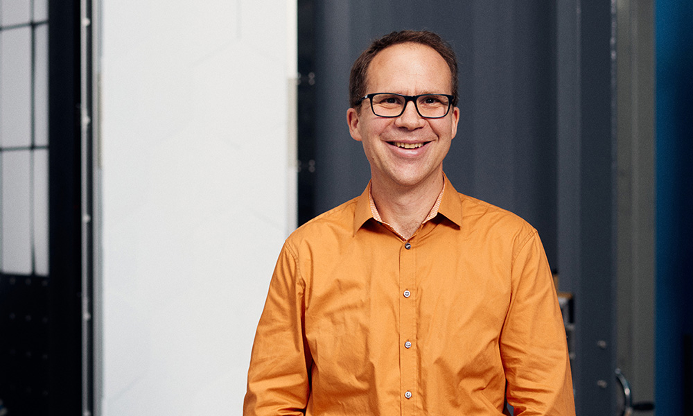 Portraitfoto von Marc Aliva, lächelnd mit orangem Hemd und Brille