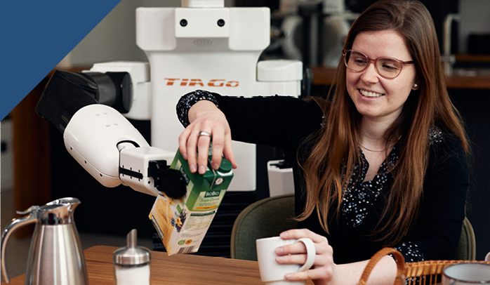 Roboter im Hintergrund unterstützt eine lächelnde jungen Frau beim Einschenken von Milch in eine Tasse.