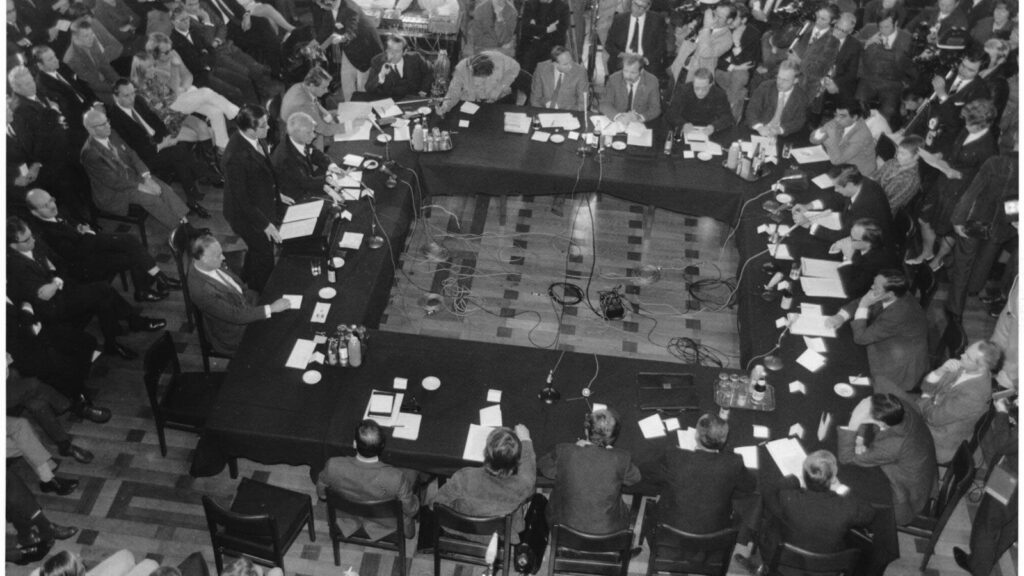 Menschen sitzen an einem großen Tisch, umringt von Menschen aus dem Publikum und Presse-Vertretern.